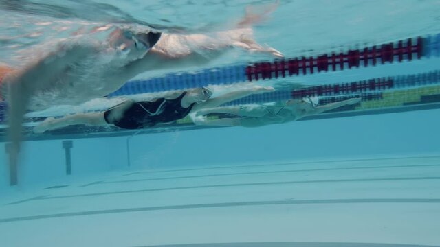 Underwater tracking shot of one Caucasian male and two Caucasian female swimmers gliding through the water and swimming in the freestyle, making synchronized arm and legs movement