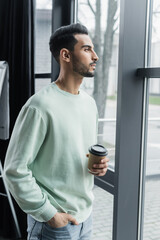 Side view of young arabian businessman holding coffee to go near window in office