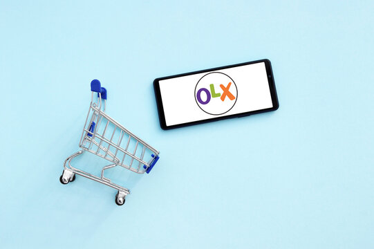 Shopping cart with a smartphone opened OLX logo on the screen on a blue background.