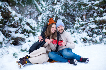 Fototapeta na wymiar A loving girl and a guy sit in the snow and drink tea from red cups in winter against the background of a snow-covered coniferous forest