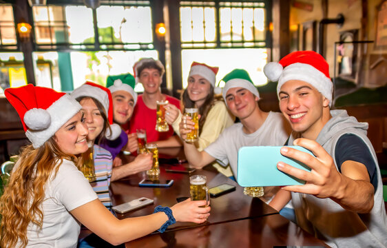 merry christmas! Young group of teenagers friends making a selfie portrait celebrating xmas time wearing santa claus hat on happy hour in irish pub drinking beer. family time concept on winter holiday