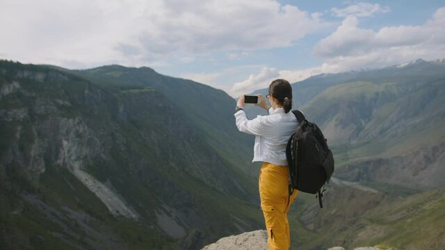 Tourist takes pictures on smartphone camera in mountains. Young woman traveler with backpack shoots video. Travel blogger on hike in mountains against backdrop of clouds