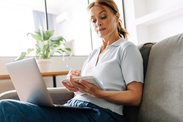 Ginger mature woman writing down notes while working with laptop at home