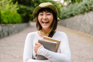 Young asian woman laughing and holding exercise books while walking at park