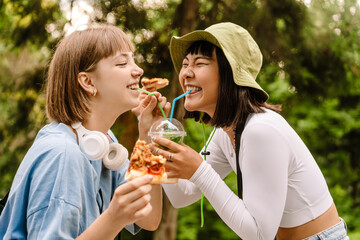 Fototapeta premium Multiracial two women drinking soda and eating pizza while resting in park