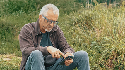 Naklejka na ściany i meble An elderly gray-haired, bearded man with glasses sits in nature and looks at the phone on a summer evening.