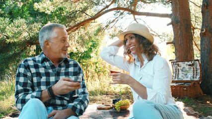 A married couple is relaxing in nature and drinking wine, and having fun talking while sitting in the woods on a picnic on a summer day.