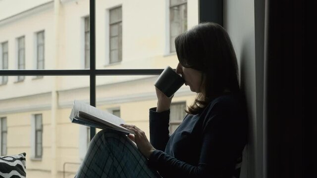 Young Female Sitting on a Windowsill with Cup of Tea or Coffee and Reading a Book. Lifestyle and People Concept