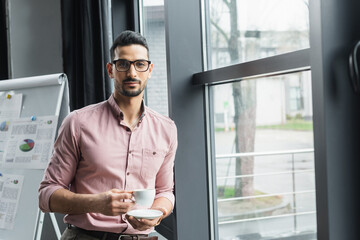 Young muslim businessman in eyeglasses holding cup and saucer in office
