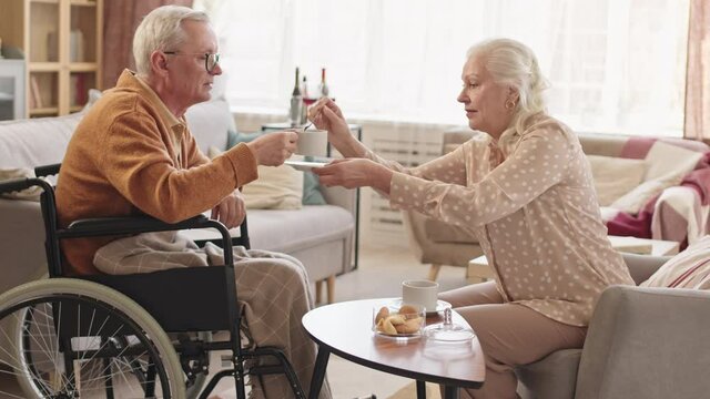 Side view medium long of older Caucasian man in wheelchair sitting in front of white-haired wife, hetero couple drinking tea and chatting at home