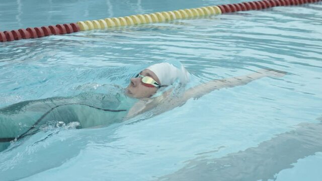 Tracking slow motion shot of a Caucasian female professional athlete swimming backstroke in the outdoor pool. The swimmer performing backstroke lying flat on the back with arms stretched