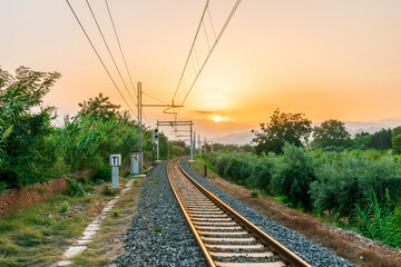 Plakat old evening railroad leading to a sunset glow in mountains with green bushes and cjlorful cloudy sku on the background