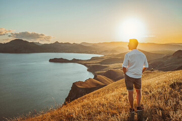 A young man travels along the mountain coast at sunset. Rear view A man stands on a mountainside...