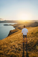 A young man travels along the mountain coast at sunset. Rear view A man stands on a mountainside and enjoys the view. The concept of freedom and achievement of goals.