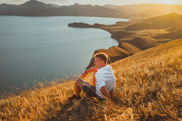 A  young man traveler sits a top a mountain in grass, enjoying the view of the coast in Crimea. Concept travel in mountains, active lifestyle and freedom  
