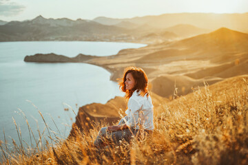 A beautiful young woman tourist is sitting on top of a mountain and enjoying the view of the sunset sea and mountains. The concept of freedom and meditation, an active lifestyle.