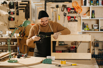Working carpenter in a watch cap busy in a big workshop. Drilling wooden piece