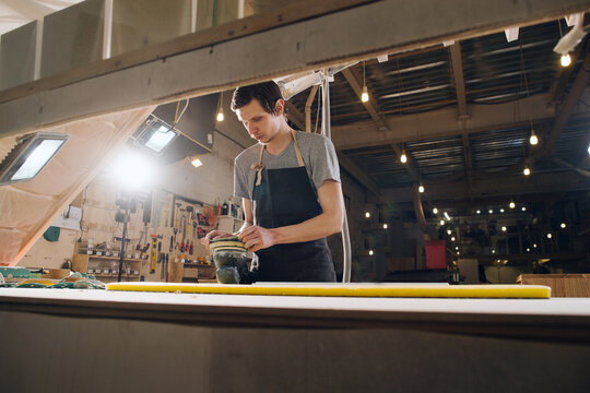 Low angle image of a craftsman grinding a wooden plank. In a big workshop.