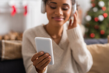 Fototapeta na wymiar Blurred african american woman in headphones using smartphone during christmas at home