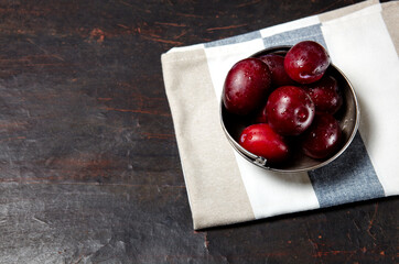Ripe red plums in bowl with water drops. Juicy fruit on wooden background, closeup. Healthy food on dark kitchen table