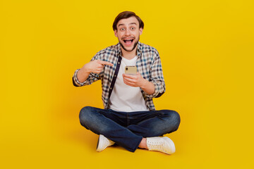 Full length photo of excited cheerful man sit floor point finger cellphone advert recommend promo isolated yellow color background