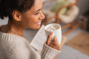 Side view of happy african american woman holding cup of cocoa with marshmallows and blurred book...