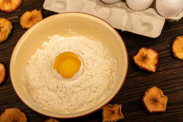 A broken white chicken egg in a bowl with wheat flour, dried apple chips and several whole eggs in a tray made of white cardboard, close-up selective focus.