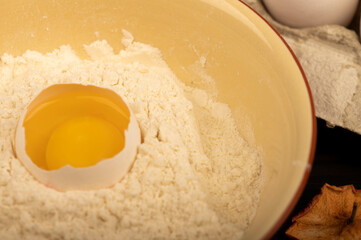 A broken white chicken egg in a bowl with wheat flour, dried apple chips and several whole eggs in a tray made of white cardboard, close-up selective focus.