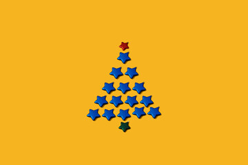 Christmas tree made of blue star shaped bauble decoration. Minimal New year concept on a yellow background. Holiday card or wallpaper pattern. Christmas spirit.