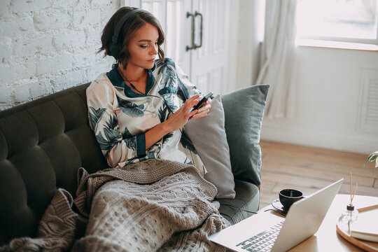 A Young Brunette Girl Works Remotely From Home Behind A Laptop In Headphones, On The Couch In Pajamas With A Warm Blanket And Uses A Mobile Phone