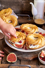 Choux rings with cream, figs and raspberries. Wooden background, side view. Children's hand holding a choux ring.