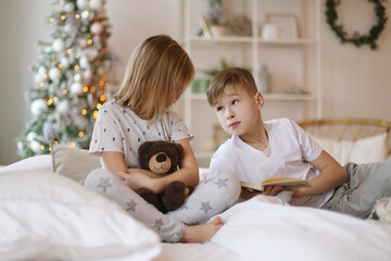 Brother is reading a book to his sister, sitting on the bed.