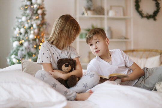 Brother is reading a book to his sister, sitting on the bed.