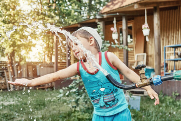 Blond little kid boy turns lever on tap with rubber hose and running up water playing in country house backyard on summer evening
