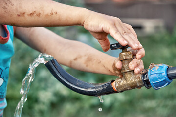 Little kid boy with dirty hands turns lever on tap with hose and running up water in country house yard in summer closeup