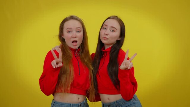 Front view two young beautiful coquette women posing at yellow background gesturing peace gesture. Happy Caucasian twin sisters looking at camera with kissing facial expression. Flirt and femininity