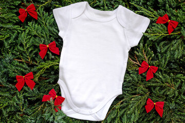 Little white cotton bodysuit for baby with copyspace lying on fir tree branches near festive red...