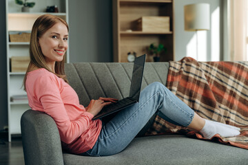 Full body profile side photo of young woman with happy positive smile sitting at the couch and browsing laptop indoors