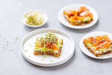 Flat lay top view of healthy sandwich with smoked salmon, cucumber, cream cheese and fresh microgreens alfalfa sprouts on white plate on gray concrete background. Healthy lifestyle. Growing sprouts