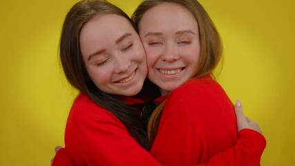 Smiling twin sisters hugging looking at camera posing at yellow background. Portrait of joyful young beautiful slim Caucasian women embracing. Unity and family concept - Powered by Adobe