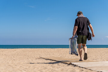 Man seen from behind walking down the wooden walkway with a beach chair near the sea on a sunny day.