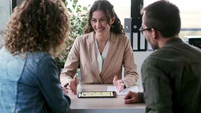 Video of beautiful real-estate agent showing house plans on electronic tablet while talking to the couple about buying the house in a real estate