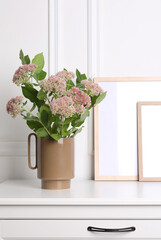 Stylish ceramic vase with beautiful flowers and blank frames on chest of drawers near white wall