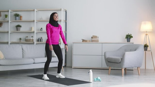 Back and buttocks training. Young active arabic lady wearing hijab practicing deep squats exercise at home, free space