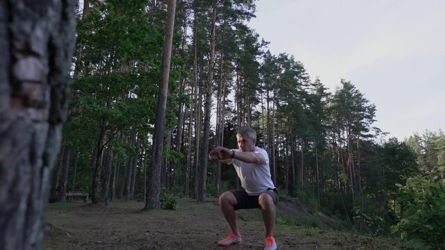 Caucasian mature person doing squat exercise with bodyweight in forest. Sporty and wellness lifestyle concept.