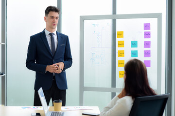 Handsome caucasian businessman are presentations of information business plans and strategy on a glass board with sticky notes to business woman are Sitting at desk with a laptop and a cup of coffee.