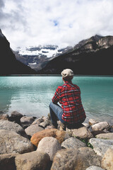 Woman in red plaid shirt sitting on rocks admiring the view of lake and mountains