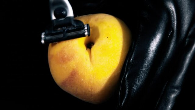 Closeup shot: shaving a peach with a razor (with the help of hands wearing black gloves). Visual representation of female or male genitals.
