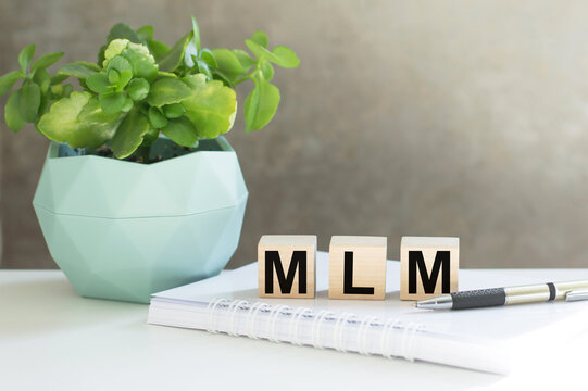 mlm - multi level marketing text on wood cubes next to a potted plant and a pen with a notebook