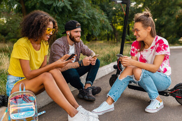 happy young company of smiling friends sitting in park on grass with electric kick scooter